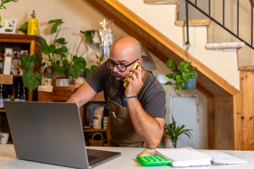 40ish bald man with glasses and a beard talking on a cell phone while looking at a laptop screen